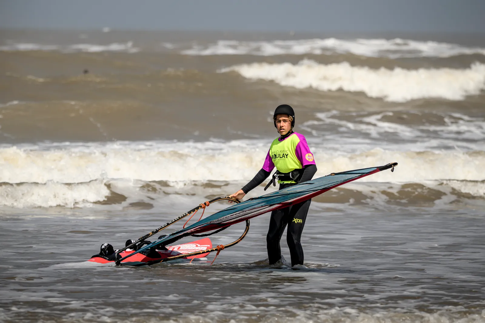 Young rider in a competition lycra walking out of the shorebreak with board and sail